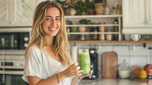 A young woman standing in a bright kitchen, holding a green smoothie with a happy smile
