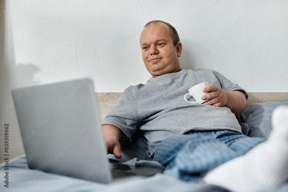 A man with inclusivity sits on a bed, relaxed and looking at a laptop. He holds a white mug in one hand and has his feet up. A man with inclusivity sits on a bed, relaxed and looking at a laptop. He holds a white mug in one hand and has his feet up.