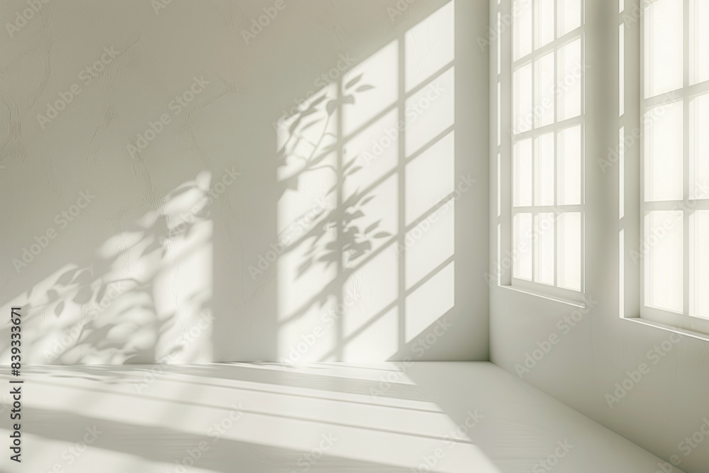 Fototapeta premium Soft sunlight casts intricate leaf shadows through large windows, creating a minimalist and serene atmosphere within an empty, white-walled room.