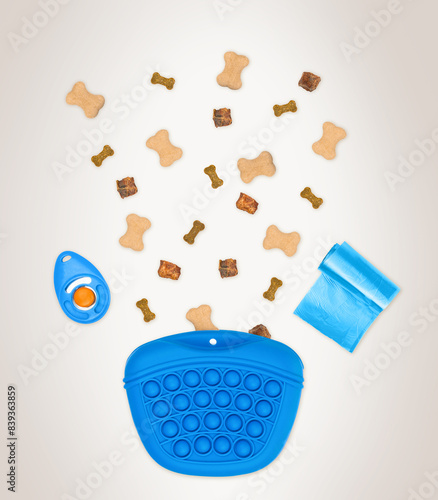 Blue dog training bag containing bone-shaped treats, a blue clicker and blue poop bags on beige background. Dog training equipment.