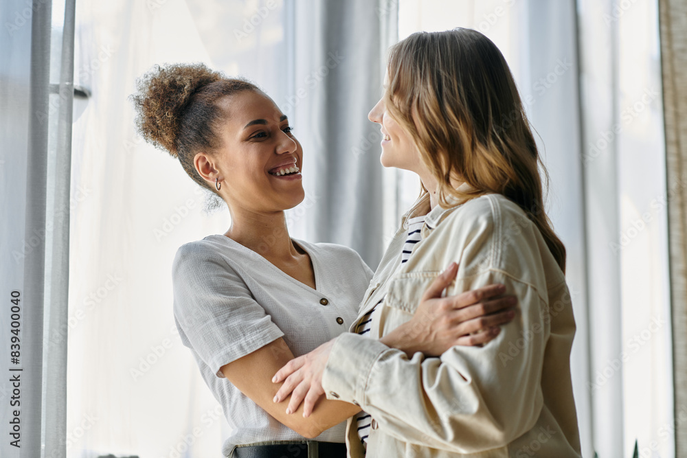 Two women, one with blonde hair and one with curly brown hair, embrace in a sunny room. Two women, one with blonde hair and one with curly brown hair, embrace in a sunny room.