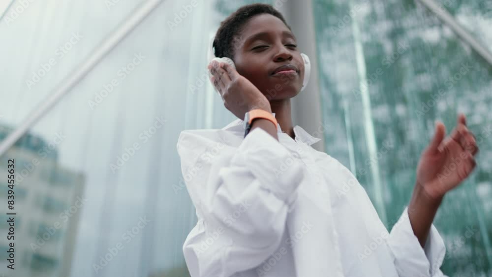 Smiling woman in wireless headphones, enjoying favorite playlist while standing in front of modern glass building. Female fell happiness and tranquility, joy of music and ease of modern technology
