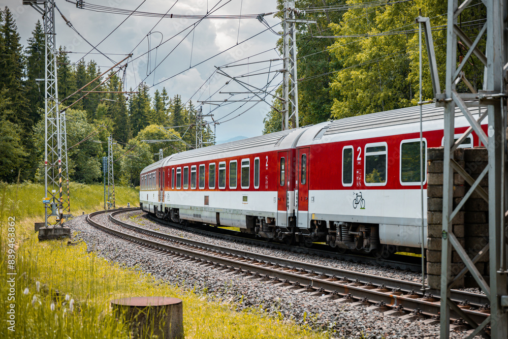 Fototapeta premium Modern electric passenger train enters the train station. Summer landscape in background. Red passenger wagons.