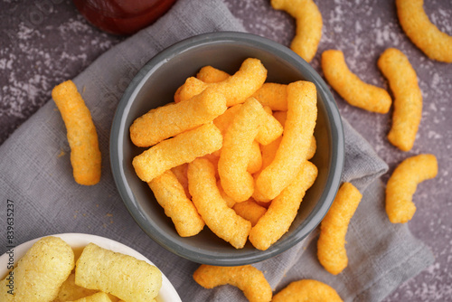 Quadro su tela Cheese puffs in bowl, crunchy corn snack close up on rustic table, top view of s
