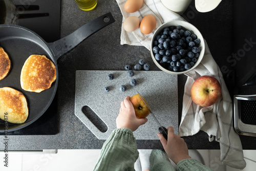 Child hands holding knife and cutting apple on stone cutting board. Various ingredients - blueberries in bowl, eggs and pancakes on griddle on kitchen counter. Preparation of healthy meal, family photo
