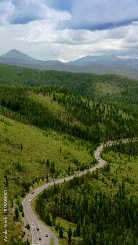 Green mountainous territories covered with fir-trees and grass. Highway in the hills nearby the mountain river. Cloudy sky at backdrop. Vertical video photo