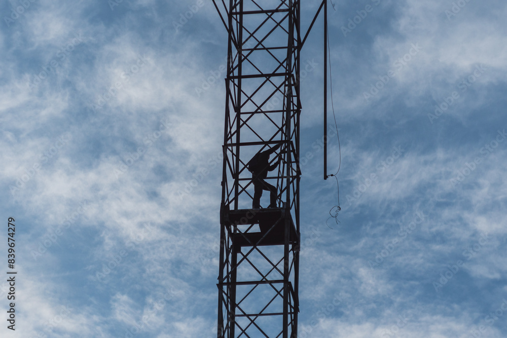 A spectacular silhouette of a man working on a metal tower against a cloudy sky. Loosely hanging cables indicate construction, repair or telecommunication work. A spectacular silhouette of a man working on a metal tower against a cloudy sky. Loosely hanging cables indicate construction, repair or telecommunication work.