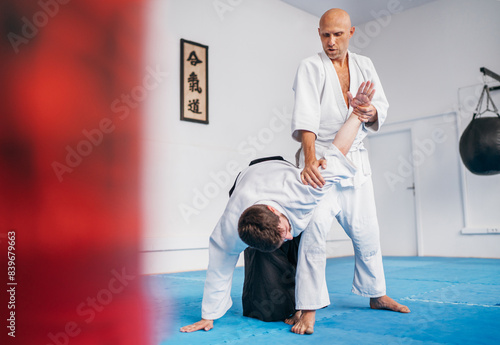 Two men train in aikido with one applying joint lock Hiji kime osae technique They are dressed in traditional martial arts kimono and Hakama, practicing under sign featuring Japanese kanji AIKIDO word photo