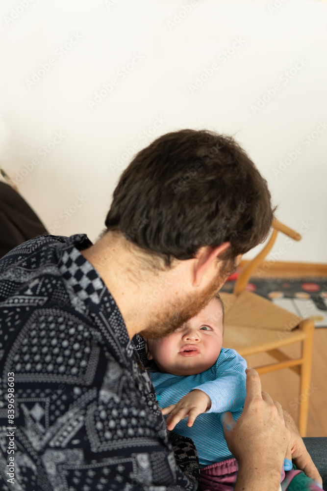 man sitting at home tries to comfort his baby dressed in a blue sweater who looks at him angrily and suspiciously. fatherhood concept, father's day man sitting at home tries to comfort his baby dressed in a blue sweater who looks at him angrily and suspiciously. fatherhood concept, father's day