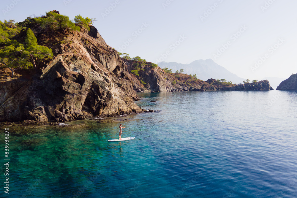 Aerial view of woman paddling a stand-up paddleboard or SUP board on a calm sea along rocky shore Aerial view of woman paddling a stand-up paddleboard or SUP board on a calm sea along rocky shore