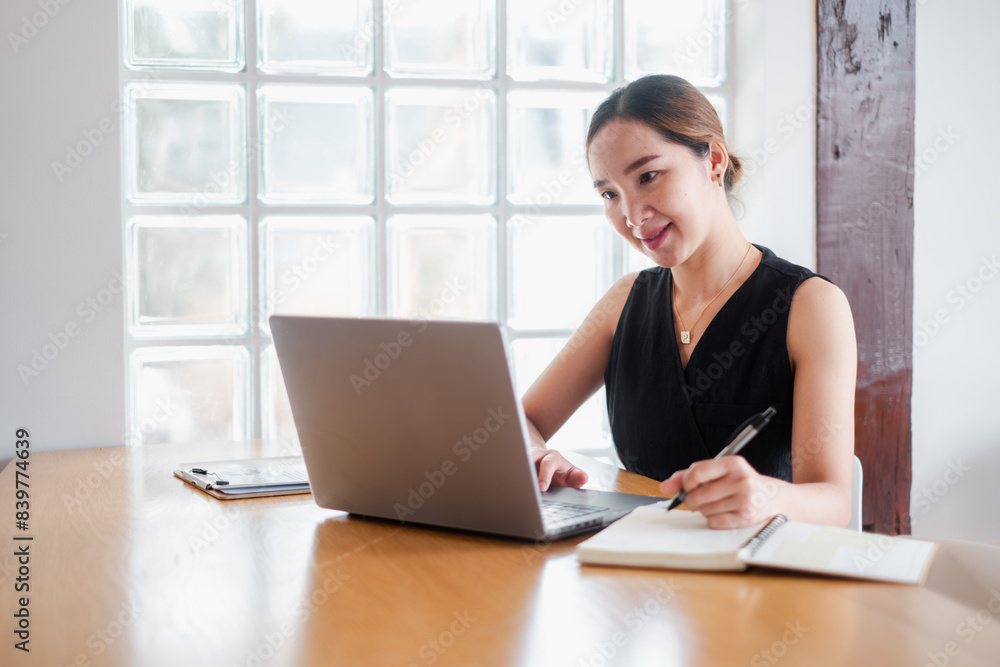 A professional woman working on a laptop and taking notes in a modern office with a glass block wall background. A professional woman working on a laptop and taking notes in a modern office with a glass block wall background.