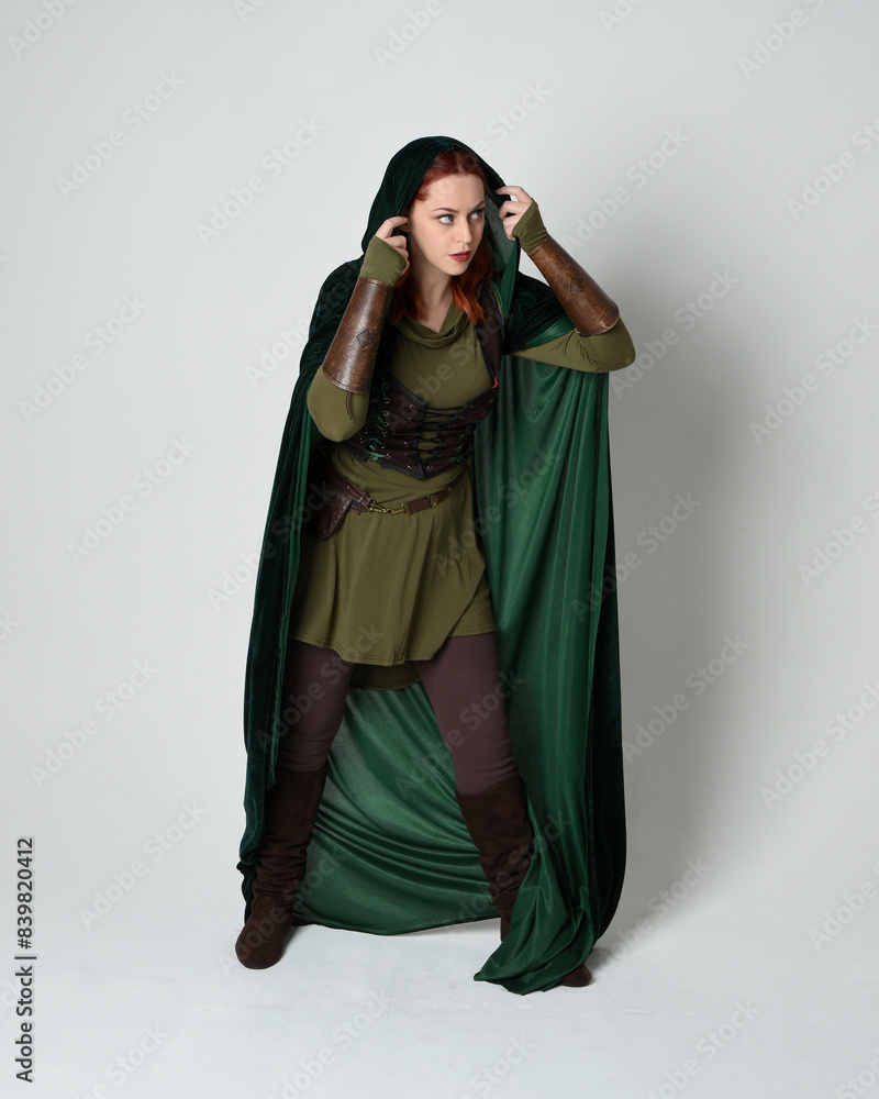 Full length portrait of red haired female model wearing green medieval fantasy costume leather armour, dramatic long flowing hooded velvet cape. Standing walking pose, isolated white studio background Full length portrait of red haired female model wearing green medieval fantasy costume leather armour, dramatic long flowing hooded velvet cape. Standing walking pose, isolated white studio background