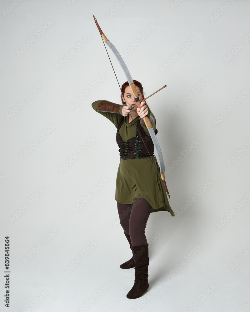 Full length portrait of red haired female model wearing green medieval fantasy costume, leather armour.
Holding archery bow and arrow weapon, standing action pose isolated on white studio background. Full length portrait of red haired female model wearing green medieval fantasy costume, leather armour.
Holding archery bow and arrow weapon, standing action pose isolated on white studio background.