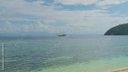 Traditional sailing boat floats in a distance on the turquoise waters of Raja Ampat, Indonesia. The serene sea creates tranquil scene of untouched beauty in tropical paradise. Static slow motion shot.