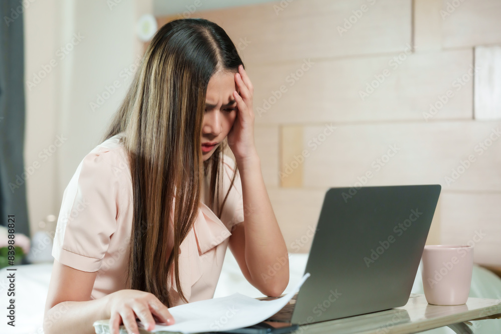Asian woman frustrated while working on laptop. Holding head, documents on table, pink coffee cup. Long hair, casual outfit. Modern home office setting, beige and white decor, natural lighting. Asian woman frustrated while working on laptop. Holding head, documents on table, pink coffee cup. Long hair, casual outfit. Modern home office setting, beige and white decor, natural lighting.