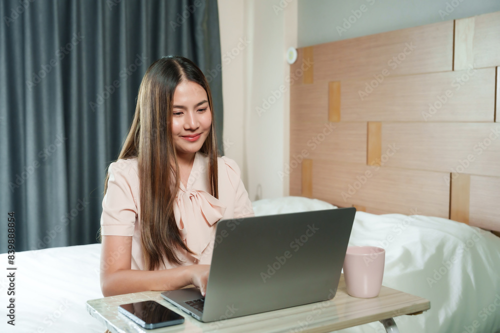Asian woman working on laptop in bedroom. Pink coffee mug on table. Long hair, casual attire, focused expression. Modern home office setup with natural light. Comfortable and cozy environment. Asian woman working on laptop in bedroom. Pink coffee mug on table. Long hair, casual attire, focused expression. Modern home office setup with natural light. Comfortable and cozy environment.