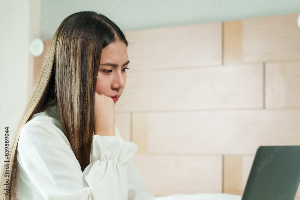Asian woman working on laptop with focused expression. Long hair, casual outfit, hand resting on chin. Modern home office setup, beige decor, natural light, indoor environment, concentrated on task. Asian woman working on laptop with focused expression. Long hair, casual outfit, hand resting on chin. Modern home office setup, beige decor, natural light, indoor environment, concentrated on task.