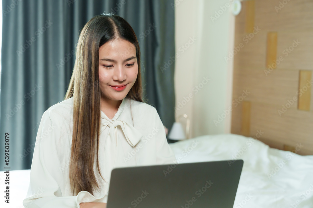 Young Asian woman smiling while working on laptop. Casual attire, modern bedroom setting. Home office setup, beige decor, natural light. Relaxed and focused expression, pink coffee cup on table. Young Asian woman smiling while working on laptop. Casual attire, modern bedroom setting. Home office setup, beige decor, natural light. Relaxed and focused expression, pink coffee cup on table.
