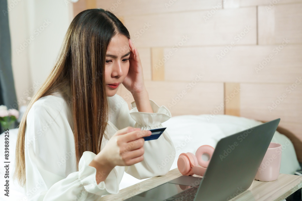 Asian woman looking concerned while holding credit card in front of laptop. Long hair, casual outfit. Modern home office setup, beige decor, natural light. Pink headphones on table, focused expression Asian woman looking concerned while holding credit card in front of laptop. Long hair, casual outfit. Modern home office setup, beige decor, natural light. Pink headphones on table, focused expression