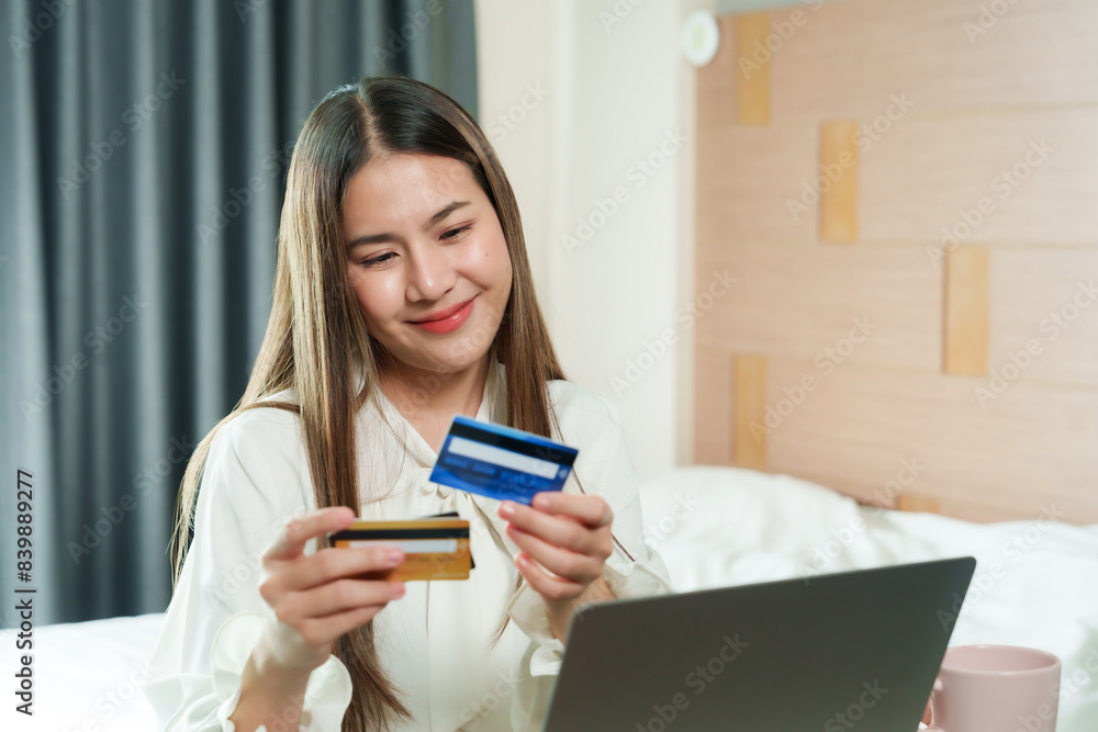 Asian woman smiling while holding credit cards in front of laptop. Long hair, casual outfit. Modern home office setup, beige decor, natural light., pink coffee cup on table, indoor environment. Asian woman smiling while holding credit cards in front of laptop. Long hair, casual outfit. Modern home office setup, beige decor, natural light., pink coffee cup on table, indoor environment.