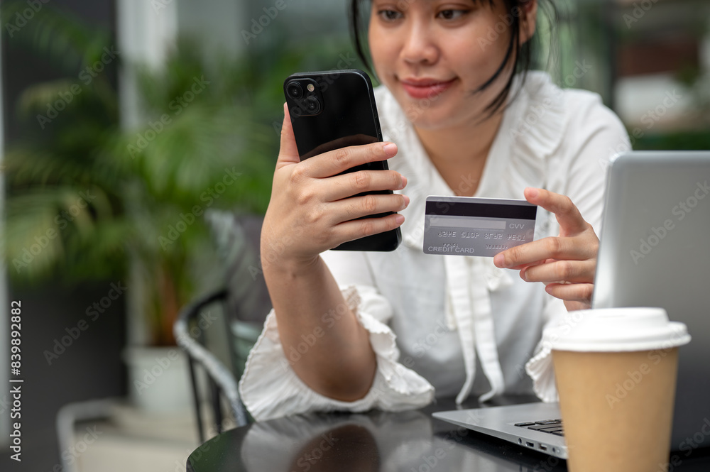 A young Asian woman holding a credit card and her smartphone while sitting at a table outdoors. A young Asian woman holding a credit card and her smartphone while sitting at a table outdoors.