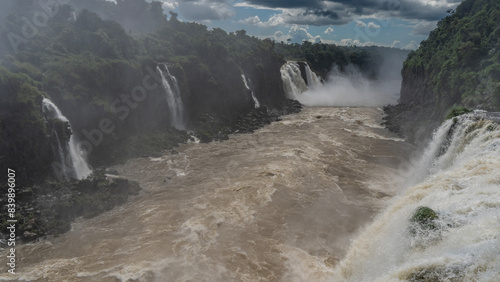 A rapid river in the gorge. The stormy water is swirling. Streams of powerful waterfalls fall from the cliffs. Splashes  fog. Green tropical vegetation. Clouds in the blue sky. Iguazu Falls. Brazil.