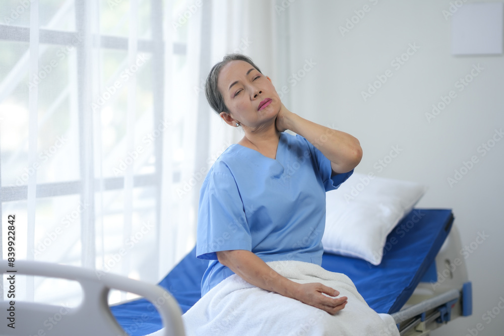 Fototapeta premium Woman in hospital gown sitting on a bed, holding her neck in discomfort, in a bright hospital room with white curtains.