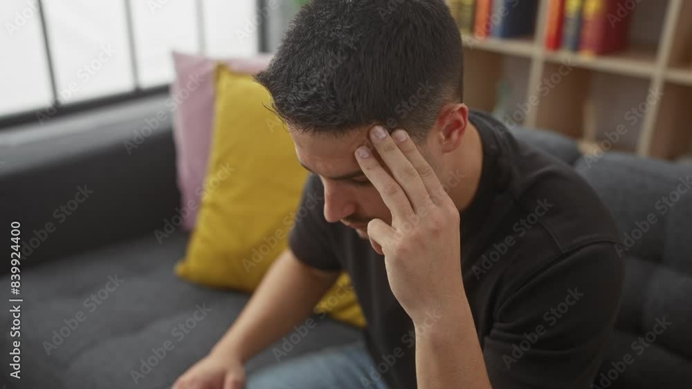A handsome young hispanic man sitting thoughtfully on a gray sofa indoors, displaying a pensive mood and casual style. A handsome young hispanic man sitting thoughtfully on a gray sofa indoors, displaying a pensive mood and casual style.