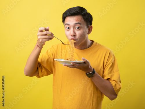 A young man in a yellow shirt is enthusiastically eating a plate of noodles with a fork against a bright yellow background.
