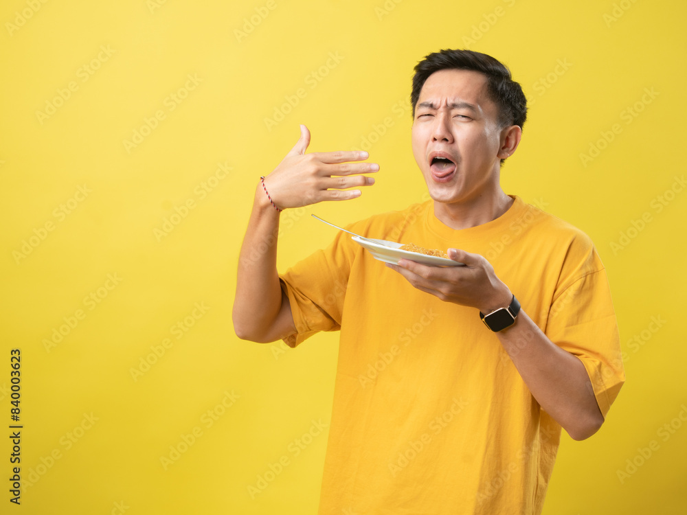 A young man in a yellow shirt reacts to hot or spicy noodles on a plate, using a fork, against a bright yellow background. A young man in a yellow shirt reacts to hot or spicy noodles on a plate, using a fork, against a bright yellow background.