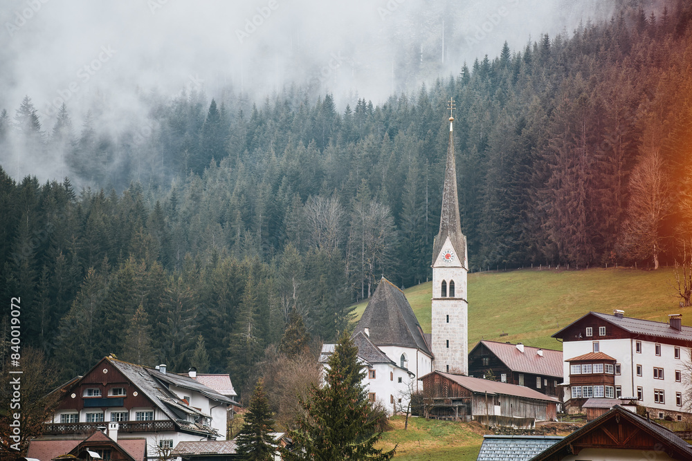 Fototapeta premium Ethereal alpine village with misty church spire among forested hills