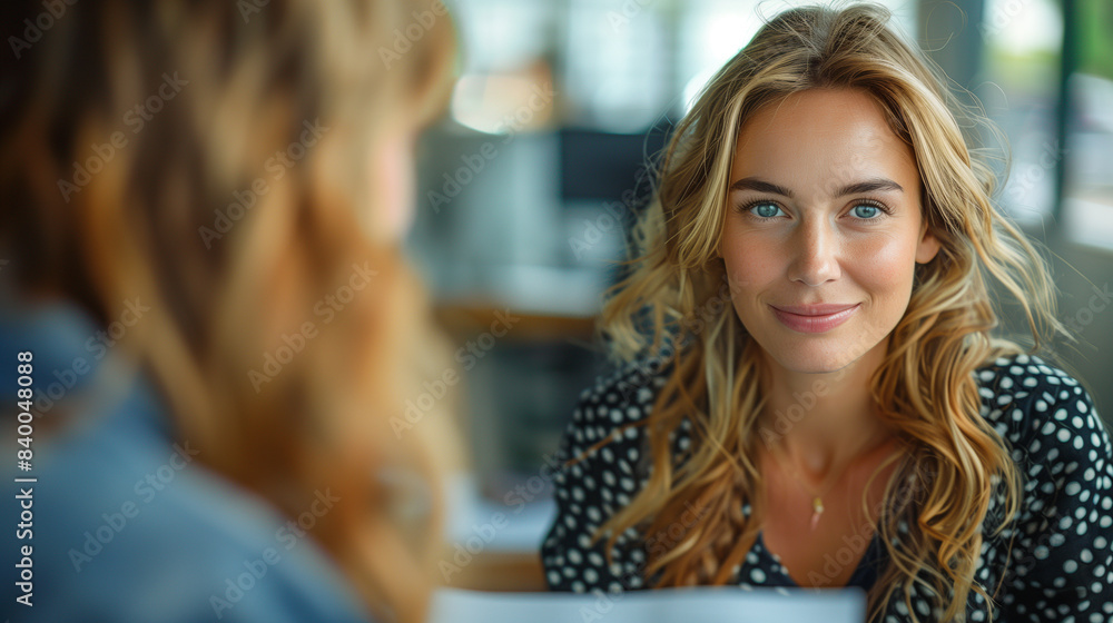 Fototapeta premium A woman with blonde hair and blue eyes smiles warmly while looking at someone off-camera in a cozy coffee shop