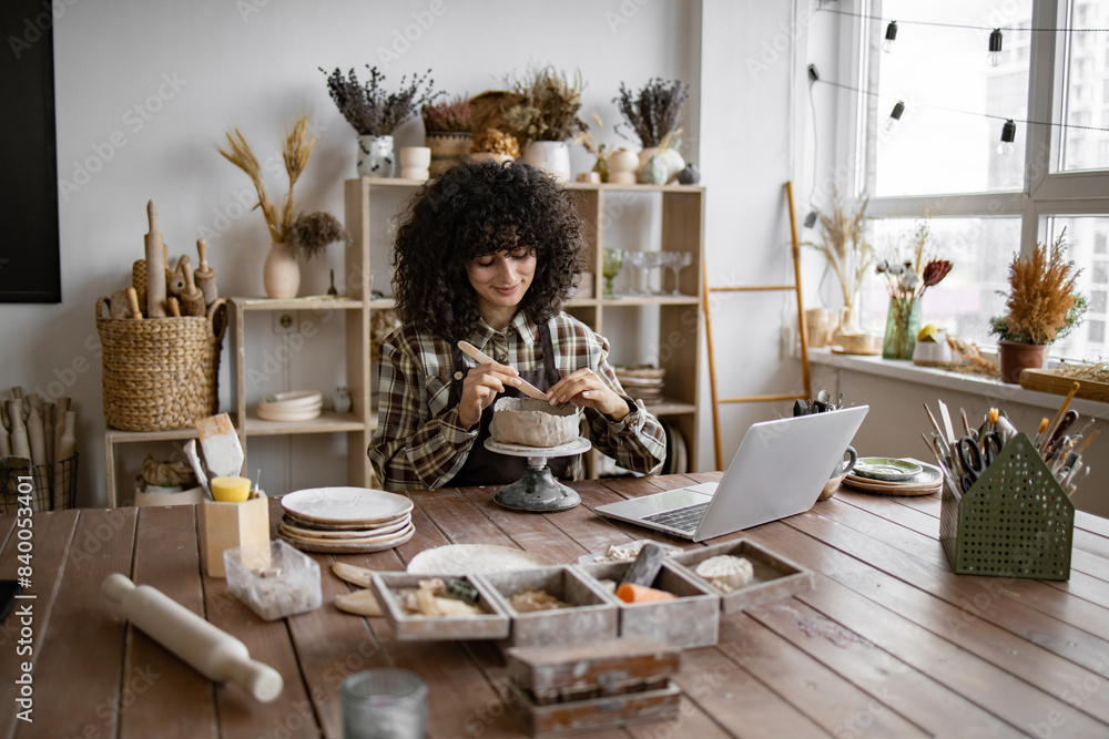 Female artist with curly hair working on pottery at wooden table in bright studio space with various artistic tools and laptop. Female artist with curly hair working on pottery at wooden table in bright studio space with various artistic tools and laptop.