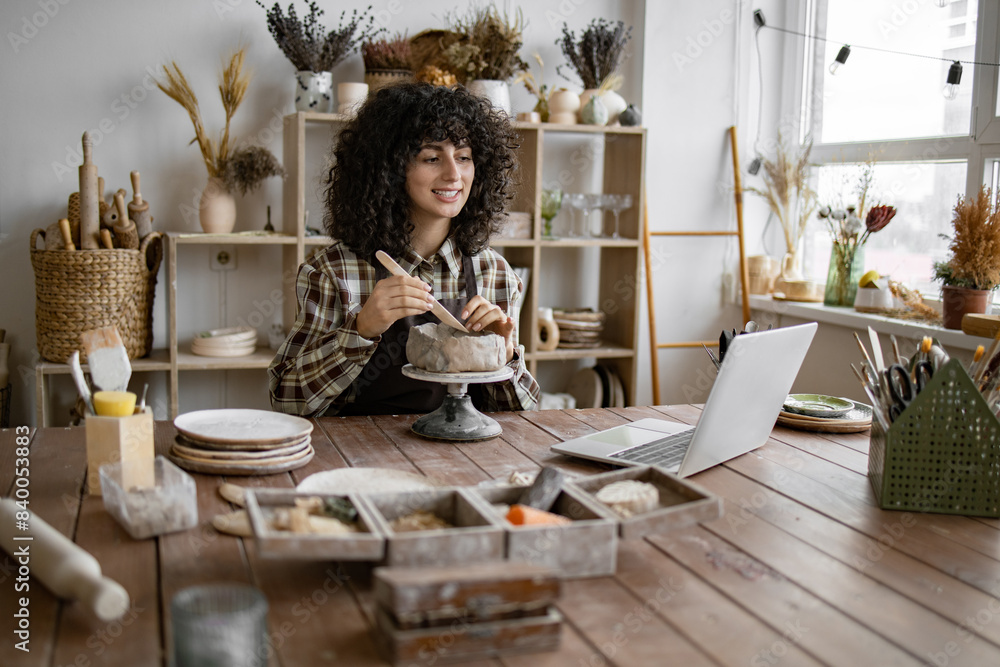 Female artist sculpting clay, smiling while video chatting in cozy, creative studio with laptop and pottery tools.