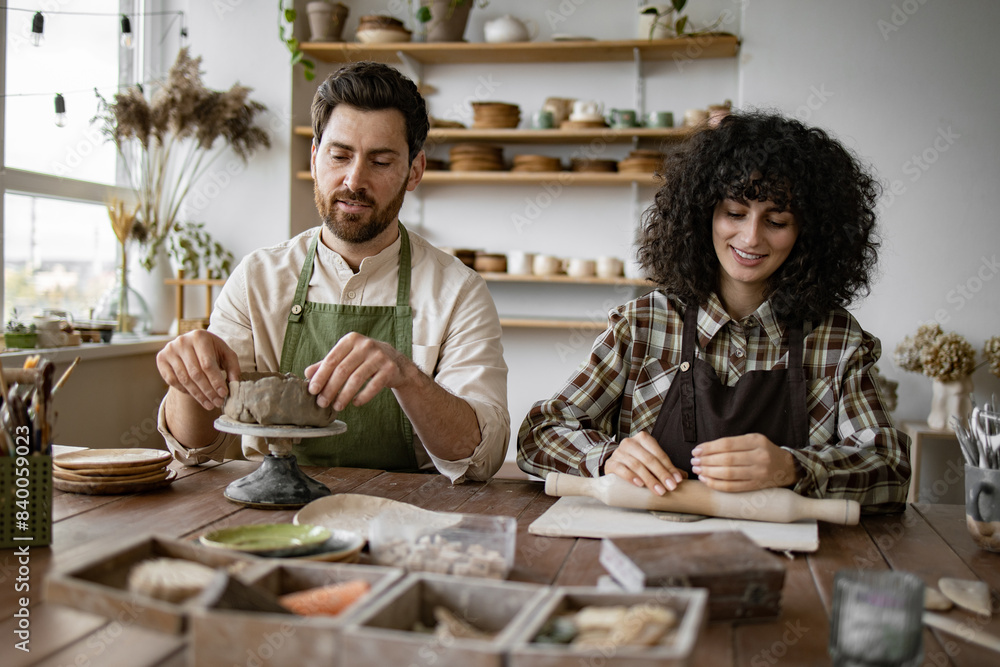 Bearded mature man and woman sculpting clay in pottery studio. Man shapes vase, woman rolls clay with rolling pin. Bearded mature man and woman sculpting clay in pottery studio. Man shapes vase, woman rolls clay with rolling pin.