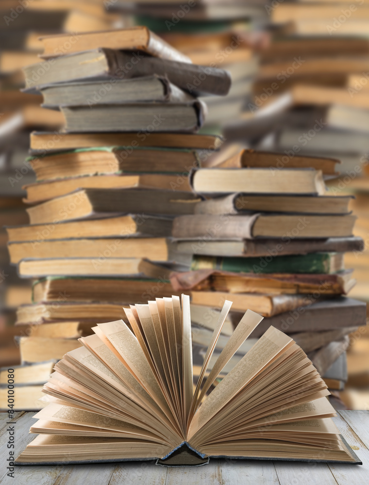 Obraz premium A single open book rests on a wooden surface, its pages fanned out in a graceful arc, against a blurred backdrop of a large stack of books.