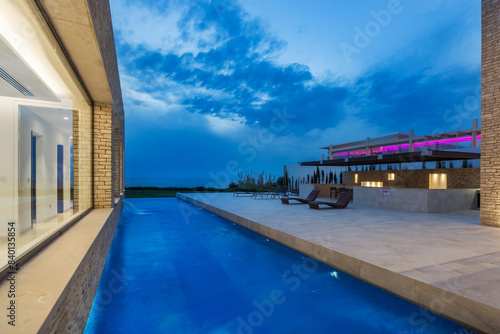 Villa with a swimming pool and a tiled deck with sun loungers against the background of the dramatic evening sky. The tranquil waters of a luxurious swimming pool reflect the vivid hues of the evening
