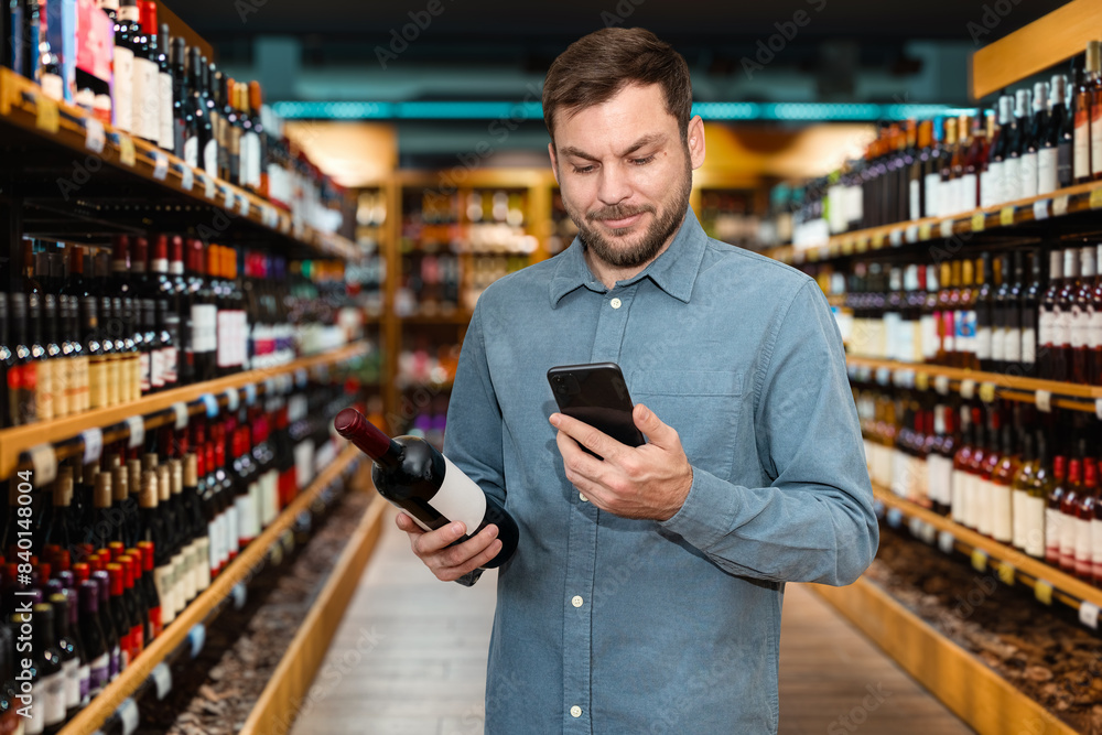 Handsome man choosing wine in liquor store. Male customer holds red wine bottle and mobile phone in his hands while shopping in wine shop. Handsome man choosing wine in liquor store. Male customer holds red wine bottle and mobile phone in his hands while shopping in wine shop.