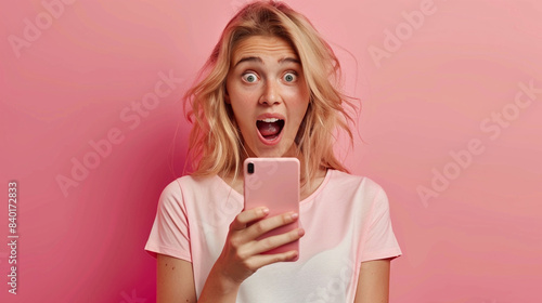 Beautiful girl looks at smart phone with surprised open mouthed expression as she wins prize, earns money, watches unbelievable post on social media, watches shocking video photo