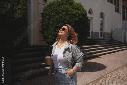 Plus size woman dressed jeans shirt, sunglasses, t-shert and skirt ealking in the street, Woman holding a cup of coffee.  The girl walking in windy day photo