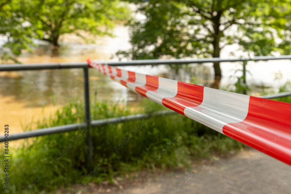 Ein rot-weißes Absperrband vor unscharfem Hintergrund mit Hochwasser, Aufgenommen in Köln, Deutschland. Ein rot-weißes Absperrband vor unscharfem Hintergrund mit Hochwasser, Aufgenommen in Köln, Deutschland.
