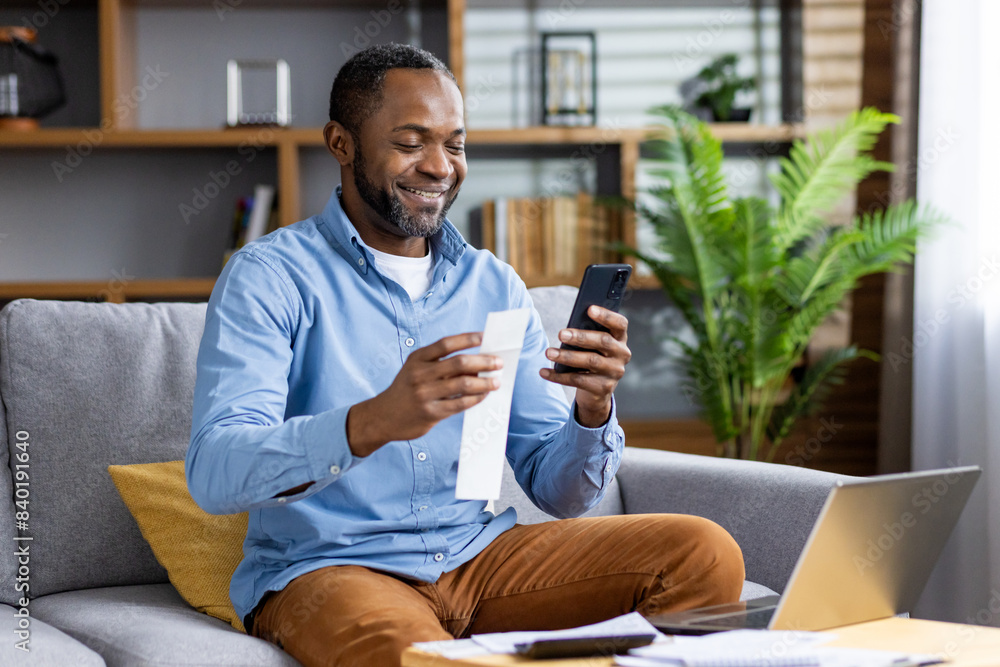 Smiling man sitting on a comfortable couch, holding a paper and using a smartphone, enjoying online services and technology in a modern home setting. Smiling man sitting on a comfortable couch, holding a paper and using a smartphone, enjoying online services and technology in a modern home setting.