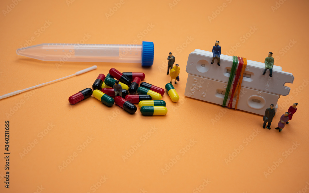 Covid-19 test cassettes (tied with rubber bands) with swab stick, drug pills, tube and human figurines; isolated on orange background. A close-up and selective focus photo of the cassette. Covid-19 test cassettes (tied with rubber bands) with swab stick, drug pills, tube and human figurines; isolated on orange background. A close-up and selective focus photo of the cassette.