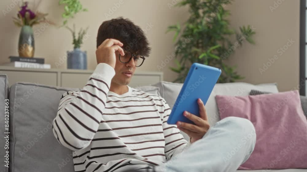 Young hispanic man wearing glasses and a striped shirt reads from a blue tablet while relaxing on a sofa in his modern living room with plants and decor, suggesting a comfortable home environment. Young hispanic man wearing glasses and a striped shirt reads from a blue tablet while relaxing on a sofa in his modern living room with plants and decor, suggesting a comfortable home environment.