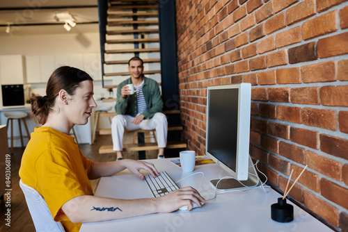 A young gay couple spends a casual afternoon in their modern apartment. One partner works at a desk while the other relaxes nearby. photo