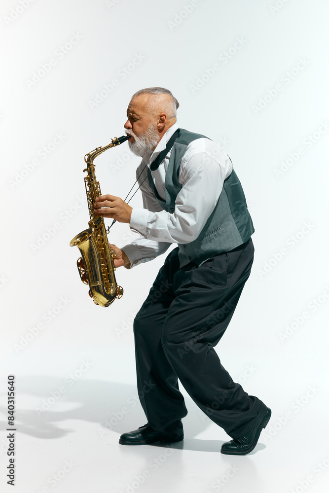 Naklejka premium Jazzy vibes. Bearded musician in sleeveless gray vest and black pants plays saxophone energetically against white studio background. Concept of music, festivals, contemporary art, hobby, creativity.
