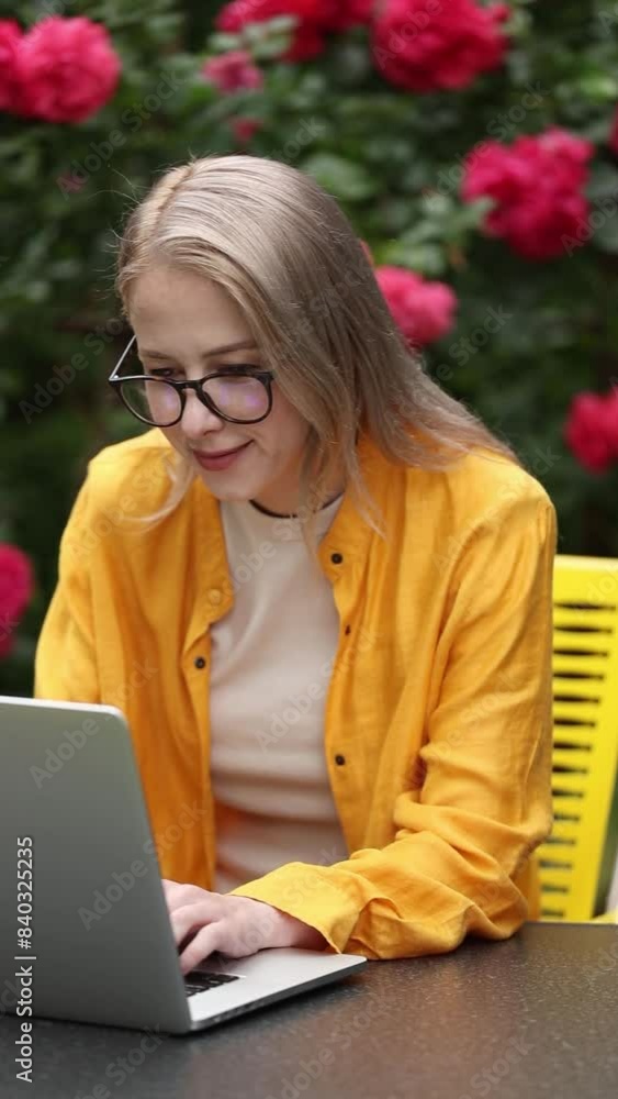 Stylish woman in yellow shirt working with laptop computer in garden with roses on background Stylish woman in yellow shirt working with laptop computer in garden with roses on background