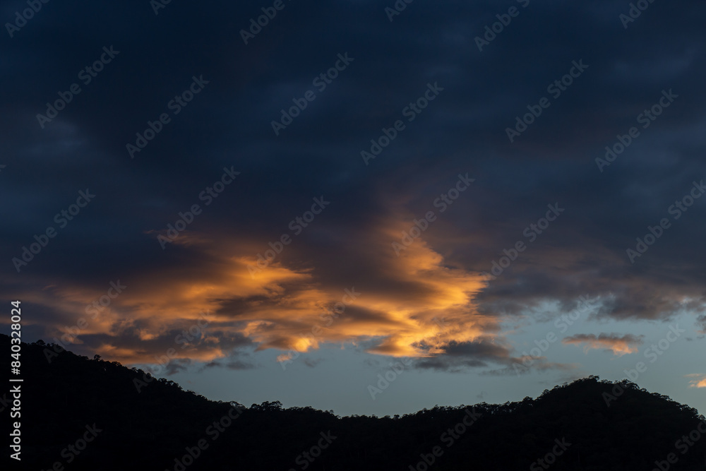 Landscape of thick clouds, orange light hitting the blue sky, silhouettes of trees, forests, and mountains. Area for text in the middle of the image Landscape of thick clouds, orange light hitting the blue sky, silhouettes of trees, forests, and mountains. Area for text in the middle of the image