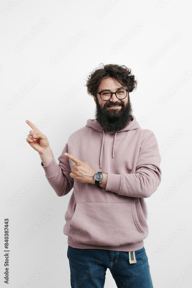 Fototapeta premium A stylish man dressed in a pink hoodie and blue jeans points emphatically to the side against a clean white background, suggesting direction, attention, or presentation.