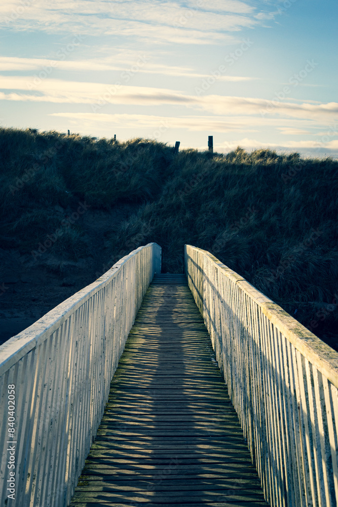 Obraz premium The footbridge over the river 'Water of Cruden', with the sun low in the sky casting a golden light over Cruden Bay Beach, Aberdeenshire, Scotland on a cold New Year's Day.